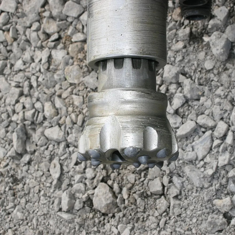 DTH hammer body with button bit fitted, resting on crushed rock at a geothermal drilling site